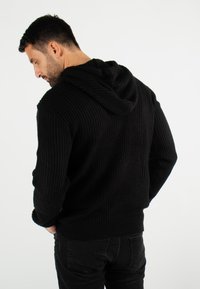 Hoodie noir en maille avec texture côtelée, capuche superposée et manches longues, associé à un pantalon foncé. Vue de dos.