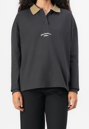 Osoba nosząca czarną koszulkę polo z długim rękawem, beżowym kołnierzykiem i białym logo Quiksilver, w połączeniu z czarnymi spodniami.