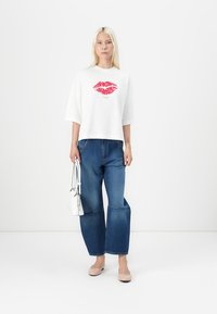 White cotton t-shirt with red lip print, paired with wide-leg blue jeans. Model holds a small white handbag and wears nude flats.