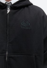 Μαύρο hoodie με φερμουάρ και κεντημένο λογότυπο "favela" στο στήθος, φορεμένο πάνω από πουκάμισο με ριγέ γιακά.