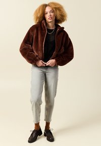 Brązowa kurtka z futra ekologicznego z wysokim kołnierzem, zamkiem błyskawicznym i obszernymi rękawami, w zestawie z jasnoszarymi dżinsami i brązowym obuwiem.