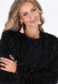 Lächelnde Frau mit langen blonden Haaren trägt einen strukturierten schwarzen Pullover, rosa Quastenohrringe und mehrere Ringe an ihrer linken Hand.