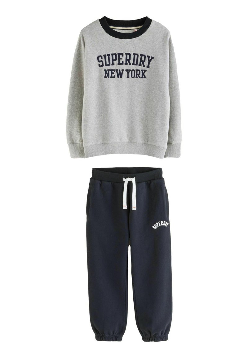 Šedá mikina s nápisom "SUPERDRY NEW YORK" v kombinácii s námorníckymi jogger nohavicami so bielym šnurovaním a logom "SUPERDRY" na stehne.