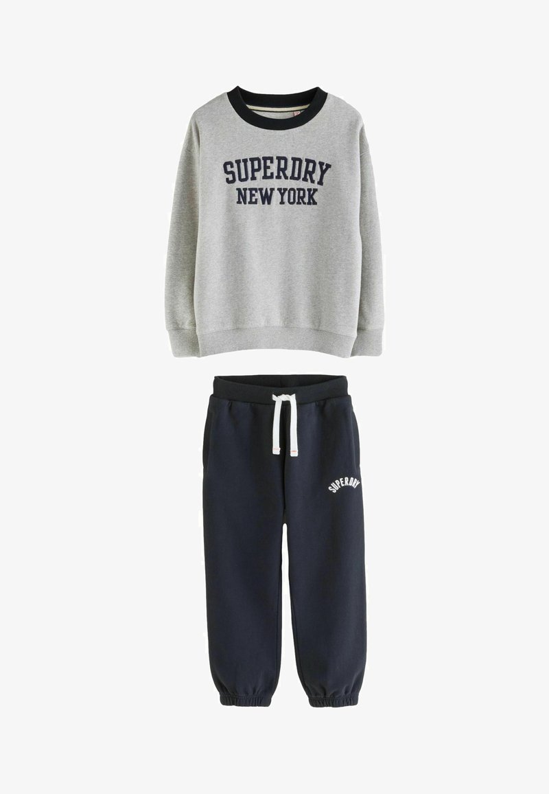 Felpa grigia con il testo "SUPERDRY NEW YORK" abbinata a pantaloni jogger blu navy con coulisse bianca e logo "SUPERDRY" sulla coscia.