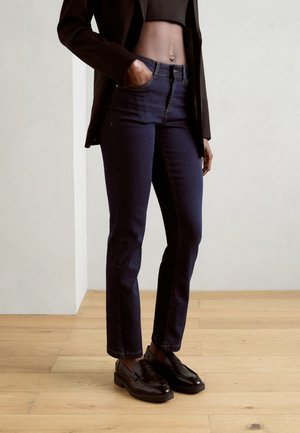 Dunkelblaue Denim-Jeans mit hoher Taille, geradem Schnitt und gelben Nähten. Dazu schwarze Loafers und ein schwarzes kurz geschnittenes Oberteil.