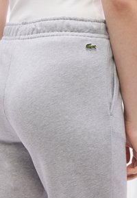 Gaiši pelēkas sporta bikses ar elastīgu jostu un sānu kabatām. Kreisajā gurnu daļā ir neliels zaļš krokodila logotips.