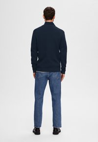 Marinblå stickad tröja med hög krage, kombinerad med blå denimjeans. Mörka skor kompletterar outfiten. Enfärgad bakgrund.