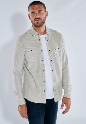 Light beige flannel shirt with vertical stripes, button-up front, two chest pockets, and black buttons. Worn over a white t-shirt.