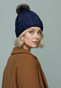 Navy knitted beanie with a ribbed design and a faux fur pom-pom on top, worn by a model with shoulder-length blonde hair.