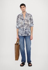 Camicia a maniche lunghe blu chiaro con fantasia floreale e bottoni, abbinata a jeans blu sbiaditi e sandali neri, mentre tiene una busta di carta marrone.