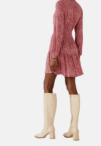 Rotes gemustertes Kleid mit langen Ärmeln und tailliertem Schnitt, kombiniert mit kniehohen beigen Stiefeln mit Absatz und seitlichem Reißverschluss.