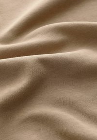 Beigefarbener Stoff mit einer weichen, glatten Textur und feinem Strickmuster, das sanfte Falten und Wellen zeigt und die Flexibilität und den Fall des Materials betont.