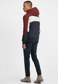 Hettejakke med fargedelt design i burgunder, krem og marineblå. Laget av et glattere stoff med elastiske ermer. Mørke jeans og grå støvler.