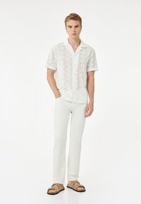 Valkoinen lyhythihainen paita, jossa on monimutkaisia leikattuja kuvioita, yhdistettynä vaaleanbeigeihin suoriin housuihin ja ruskeisiin liukuvaunupolkusaappaisiin.