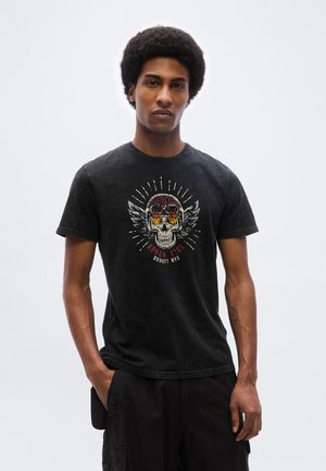 Jeune homme avec une coiffure afro portant un t-shirt noir à imprimé graphique représentant un crâne enflammé avec des ailes et un pantalon cargo noir, les mains dans les poches.