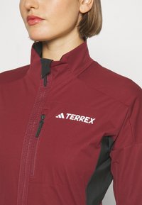 Bordeauxfarbene Terrex-Jacke mit hohem Kragen, schwarzen Akzenten, Frontreißverschluss und Logo-Detail in Weiß. Glatte Textur mit tailliertem Design.