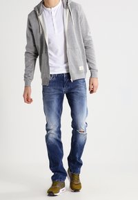 Hombre vestido con una sudadera gris con cremallera, camiseta blanca tipo henley, pantalones vaqueros desgastados azules y zapatillas de color verde oliva, de pie sobre un fondo blanco.