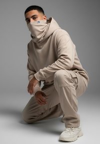 Beige hoodie with a large cowl neck, paired with matching cargo pants. Shoes are light-colored sneakers, with textured soles.