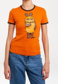 T-shirt orange avec des bordures marron présentant un design de personnage de dessin animé. Tissu doux et lisse, forme ajustée, manches courtes. Jean en denim porté en dessous.