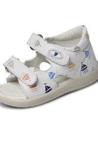 Witte sandaal met marineblauwe, lichtblauwe en beige zeilbootpatronen. Voorzien van klittenbandsluitingen en uitsparingen aan de zijkanten. Getextureerde zool.