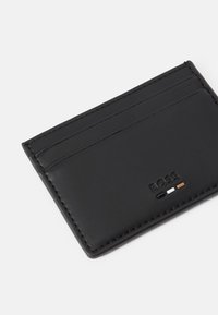 Portafoglio in pelle nera con più scomparti per carte, design elegante e cuciture a contrasto. Presenta un logo impresso e accenti di colore sottili.