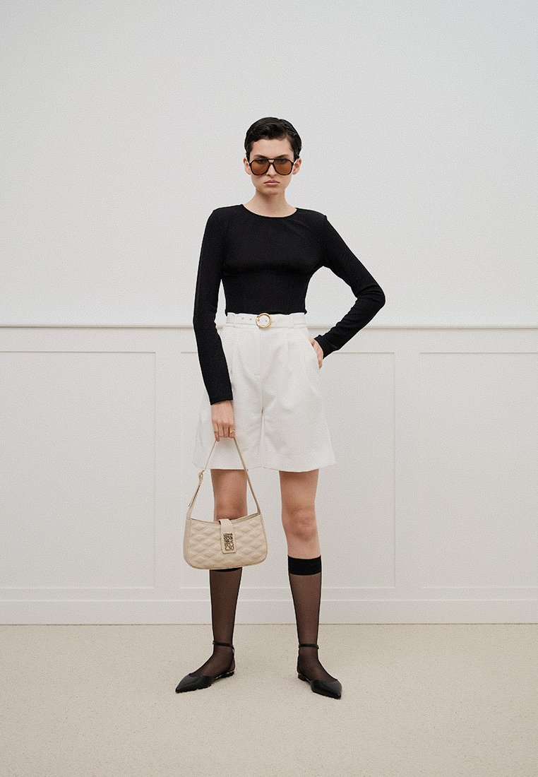 Frau steht mit der Hand auf der Hüfte, trägt ein schwarzes langärmliges Oberteil, weiße Shorts mit Gürtel, durchsichtige schwarze Socken, schwarze Ballerinas, Sonnenbrille und hält eine beigefarbene gesteppte Handtasche.