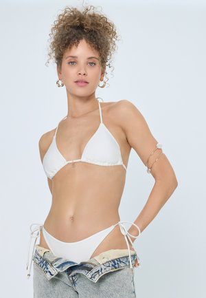 Kobieta o kręconych włosach, ubrana w biały top i dół bikini, częściowo opuszczone dżinsy, złote kolczyki-koła oraz biżuterię na ramieniu.
