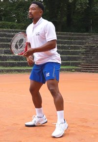 Férfi teniszjátékos egy salakpályán, fehér pólót visel logóval, kék sortot és fehér sportcipőt. Piros teniszütőt tart a kezében.