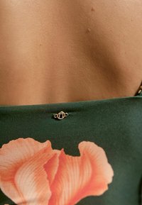 Grüner Stoff mit einem großen pinken Blumenmuster und einem kleinen goldenen Akzentdetail am Ausschnitt. Glatte Textur und taillierte Passform.
