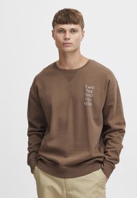 Sudadera marrón de tela suave, con cuello redondo, puños de canalé y texto bordado que dice "TÓMATE EL TIEMPO QUE NECESITES" en el lado izquierdo.
