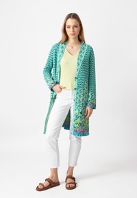 Pitkä turkoosi kardigan, jossa on geometrisiä kuvioita ja kirkas kukkakoriste hihoissa. Yllä vaaleanvihreä paita ja valkoiset housut.