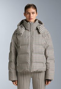 Pikowany puchowy płaszcz w brązowo-kremowy wzór w karo, z wysokim kołnierzem, zapięciem na zamek i oversized, wypchanymi rękawami.