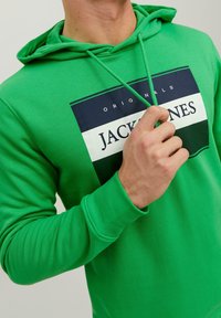 Muž v jasně zelené mikině s logem "Originals JACK & JONES", který drží jeden z tkaniček mikiny poblíž hrudi.