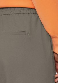 Grijze joggingsbroek met een gladde textuur, voorzien van een zijzak en een klein logo op de achterkant. Een oranje top is gedeeltelijk zichtbaar.