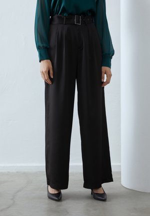 Vrouw die zwarte, wijde, hooggetailleerde broek met riem draagt, een doorschijnende teal blouse met lange mouwen en zwarte pumps met puntige neus, binnenshuis staand.