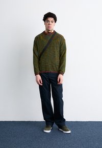 Green knitted sweater with multicolored flecks, black pants, and olive green shoes. Features a round neck and a crossbody strap.