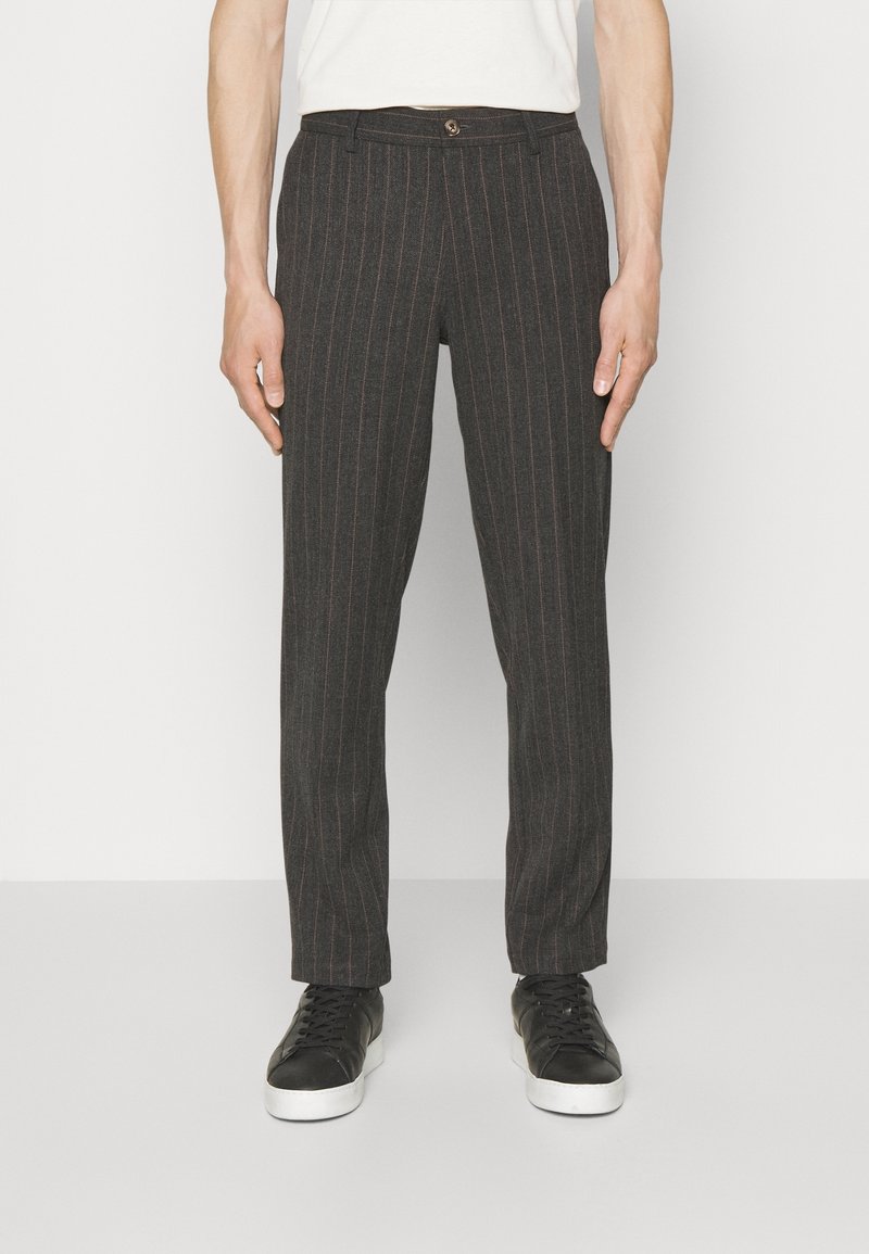 Homme portant un pantalon gris foncé à rayures fines avec fermeture par boutons et des baskets noires à semelles blanches, debout devant un fond blanc.