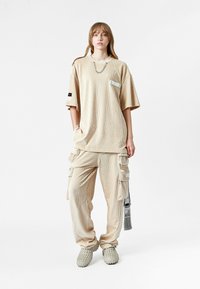 Camiseta de manga corta de punto acanalado en color beige y corte oversized, con bolsillo frontal y detalle de cadena; combinada con pantalones cargo a juego que cuentan con múltiples bolsillos y etiquetas.