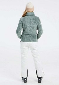 Pullover in pile verde con colletto alto, abbinato a pantaloni da sci bianchi con zip laterali, indossato con stivali da sci neri e un cappello beige.