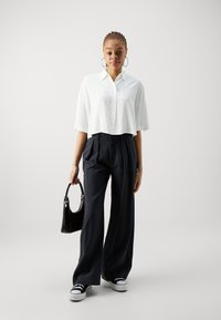 Camicia bianca a maniche lunghe, pantaloni larghi blu navy, borsa nera e sneakers nere; modello con capelli intrecciati e orecchini a cerchio.