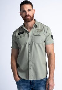 Olijfgroene shirt met korte mouwen, voorzien van knoopzakken, een kraag en geborduurde branding op de borst en mouw. Er worden denim jeans gedragen.