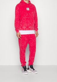 Červená mikina s batikovým vzorem, přední kapsou a nášivkou s logem, v kombinaci s odpovídajícími tepláky. Černé a šedé tenisky s bílými podrážkami.