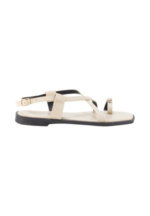 Sandalia plana beige con suela negra, con finas tiras, hebilla en el tobillo y una trabilla para el dedo con una pequeña cuenta decorativa.