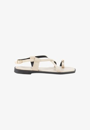 Sandalia plana beige con suela negra, con finas tiras, hebilla en el tobillo y una trabilla para el dedo con una pequeña cuenta decorativa.