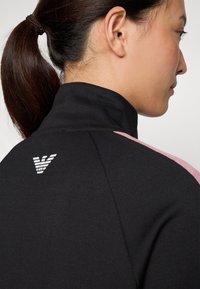 Zwarte sportjack met een hoge kraag, voorzien van roze accenten op de schouders en een wit logo op de achterkant. Glad, rekbaar materiaal.