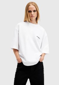 Camiseta de algodón blanca de corte oversize con cuello redondo y mangas cortas, que presenta un pequeño logo negro en la parte izquierda del pecho.