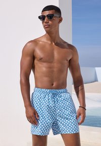 Pantalones cortos de baño azules con un patrón de huellas de manos blancas, con cintura elástica y cordón. Se llevan con gafas de sol oscuras.
