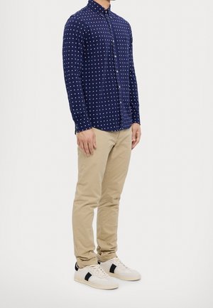 Homme portant une chemise à manches longues bleu marine à motifs, pantalon beige et baskets blanches avec des rayures noires, debout devant un fond blanc.