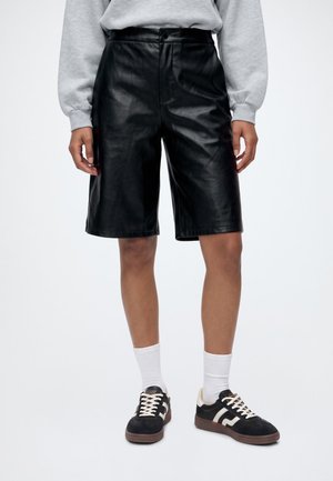 Personne portant un short en cuir noir longueur genoux, un sweat-shirt gris, des chaussettes blanches et des baskets noires avec des détails blancs.