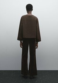 Pull texturé marron avec manches larges, associé à un pantalon marron évasé. Le modèle est de dos, face à un fond neutre.